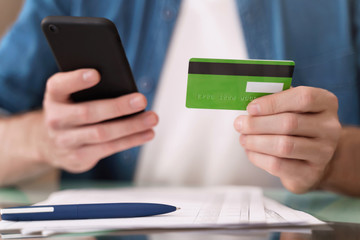 Young caucasian man paying bills online with smartphone at home. Closeup man's hands holding mobile phone and credit card. E-commerce concept