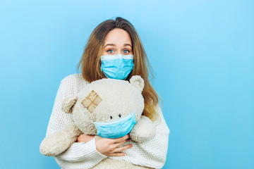 A girl in a medical mask holds a toy dressed in a medical mask on a blue background. The girl is in quarantine. Self-isolation. Coronavirus. Epidemic covid 19