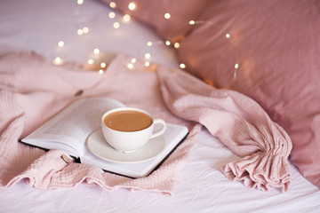 Tasty cup of coffee staying on paper book in bed over glowing lights closeup. Good morning. Breakfast.