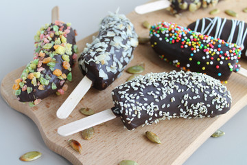 Frozen bananas covered with dark chocolate, sesame, almonds and candied fruits on wooden board....