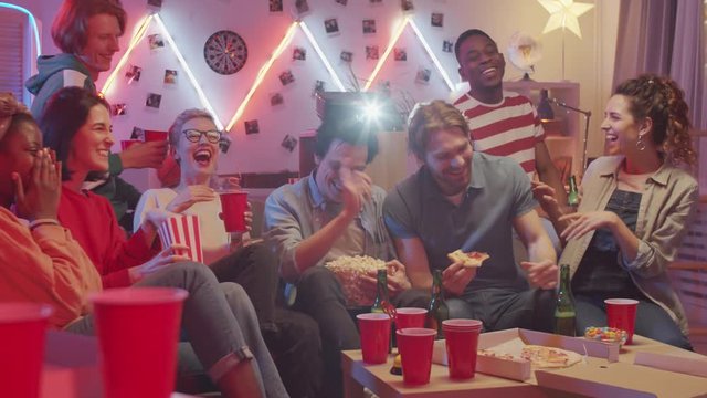 Company of multiethnic young friends sitting on couch, drinking beer, eating pizza and laughing while watching comedy show on TV at home party