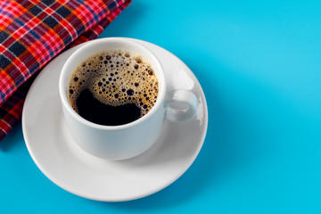 cup of coffee on a white saucer and a red checkered napkin on a blue background. foam in coffee in the form of a smile.