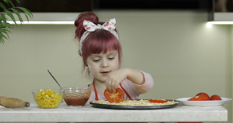 Cooking pizza. Little child in apron adding sliced tomatoes to dough in kitchen
