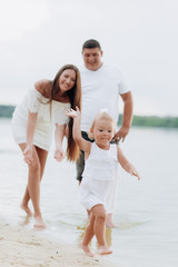 Mom, dad hugging daughter walking on the beach near lake. The concept of summer holiday. Mother's, father's, baby's day. Family spending time together on nature. Family look. Sun light.