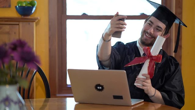 Young man in graduation costume in front of laptop in a home, taking a selfie.