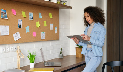 Smiling africanteen girl college student studying from home checking homework in notebook, elearning, stand at home office desk, making notes. Young black woman remote learning remote school lessons.