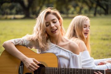 Portrait of caucasian young women sitting in the park outdoor and playing a guitar sing a song...
