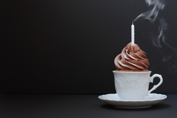 A chocolate cupcake in a small ceramic white coffee cup with a extinguished smoking candle. Black background