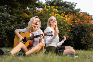Portrait of caucasian young women sitting in the park outdoor and playing a guitar sing a song together with happiness