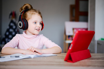 Naklejka premium Cute little girl in headphones is using a tablet while doing homework and smiling, distance education, online learning, concept