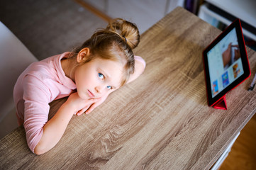 Young girl watches cartoons at home at a table on a digital tablet, a child plays with a device, a laptop computer