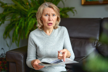 Portrait of a concerned woman reading bad news in a newspaper sitting on a couch in the living room...