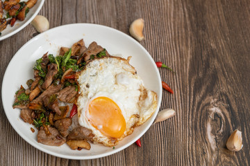 Pad Kra Pao Beef, (Stir fried beef with thai basil and chilli) top up with sun rise side up eggs on the white dish. close up , wooden table