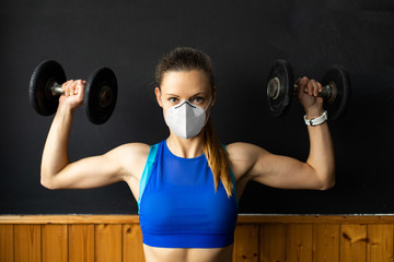 Young fit woman with n96 face mask  doing shoulder press exersice at the gym with dumbbells. Fitness strength workout under coronavirus health crisis.