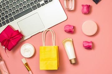 Concept of online shopping cosmetics. Top view on cosmetics bottles, cream, soap, makeup brushes, computer, paper bag on a pink background, flat lay, copy space.Layout for online cosmetics store