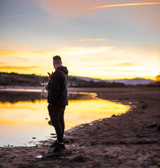 young man standing on the sand holding a dog beside a beautiful lake in the golden hour with the orange reflection of the sun in the water