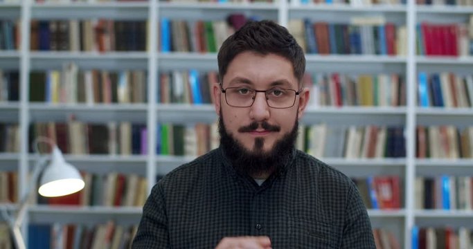 Portrait of Caucasian young man with beard standing in library room, taking on glasses and looking to camera. Male senior teacher smiling slight. Books shelves behind. Professor in bibliotheca.