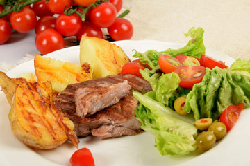 pieces of grilled steak next to potatoes grilled next to lettuce with tomatoes and olives on a white plate