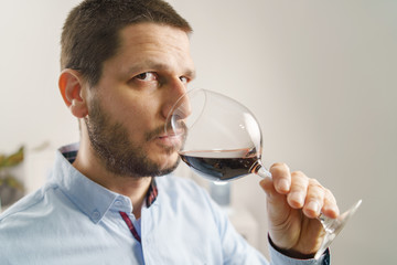 Side view portrait of adult caucasian man wearing shirt holding glass of red wine tasting at home at evening or night close up male with beard and short hair at home alone one