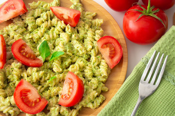 Basil pesto fusili pasta with Parmesan cheese and sliced tomatoes in a wooden plate and a green napkin with a fork in the background