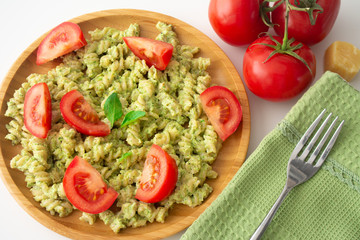 Basil pesto fusili pasta with Parmesan cheese and sliced tomatoes in a wooden plate and a green napkin with a fork in the background