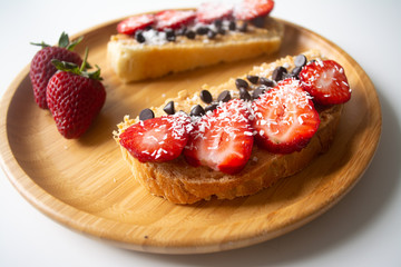 Peanut butter spread toasts with chocolate chips, strawberries, coconut flakes and homemade bread on a wooden plate