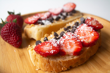 Peanut butter spread toasts with chocolate chips, strawberries, coconut flakes and homemade bread on a wooden plate