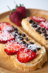 Peanut butter spread toasts with chocolate chips, strawberries, coconut flakes and homemade bread on a wooden plate