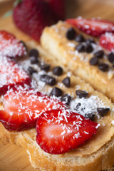 Peanut butter spread toasts with chocolate chips, strawberries, coconut flakes and homemade bread on a wooden plate