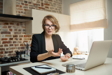 A young woman in glasses works remotely on a laptop in her kitchen. A blond girl gesticulating discusses with her colleagues on a video conference at home. A lady teaching a lesson online on a webinar