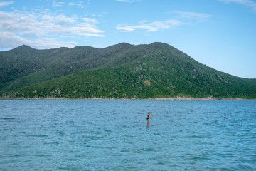 A man on a sup surfer swims on the blue water in the ocean against the background of green mountains and blue sky.