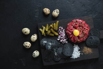 Fototapeta na wymiar Black wooden serving board with beefsteak tartare, flatlay on a black stone background, horizontal shot with space