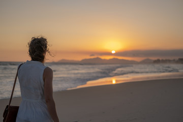 Girl in a light dress at sunset on the beach against the background of the ocean mountains and sun