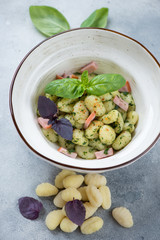 Bowl of gnocchi with green basil pesto, sliced bacon and fresh basil leaves, vertical shot on a light-blue stone surface