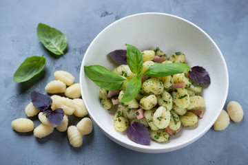 Potato gnocchi served with basil pesto sauce and chopped bacon in a white bowl, studio shot on a grey concrete background