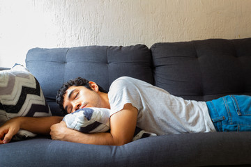 Young Hispanic man with glasses resting on a sofa in the morning, wearing jeans and a white shirt