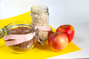 Two glass jars with buckwheat and oatmeal and two red apples on a yellow background. The concept of...
