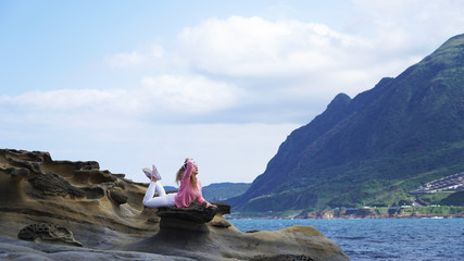 flexible European girl on stone in an unusual pose performs split in a beautiful place on the ocean with mountain and sky views, white clouds, coast, a cute curly woman traveler on a beach in Taiwan