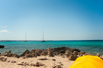 Natural and wild beach facing the sea with yellow umbrellas. Sandy and rocky beach with yellow umbrellas, in the background the Mediterranean sea and sailing boats moored. Formentera Spain
