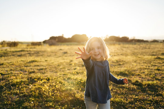 Cute little girl in casual wear with outstretched arm looking at camera with smile and showing five while standing on green meadow during summer sunset