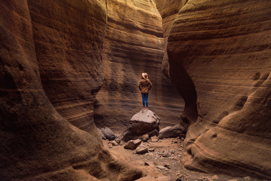 back view of woman traveler in casual outfit and hat looking up while standing on stone in deep rocky ravine with sunlight