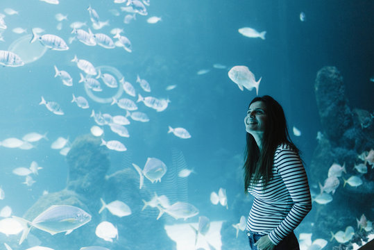 Side view of joyful black haired young woman in casual wear smiling and looking up with bright modern aquarium on background