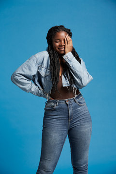 Positive beautiful African American woman with braids in denim outfit smiling with closed eyes against blue background