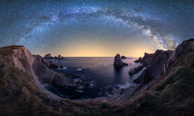 Big rough cliffs on blue calm ocean during bright evening under colorful starry sky with milky way