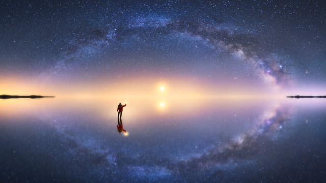 Fototapeta Silhouette of anonymous man standing on reflection surface of water and reaching out to starry colorful night sky with milky way