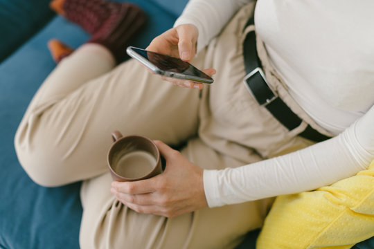 Faceless copped woman without shoes chilling on cozy sofa in office enjoying cup of coffee and surfing mobile phone