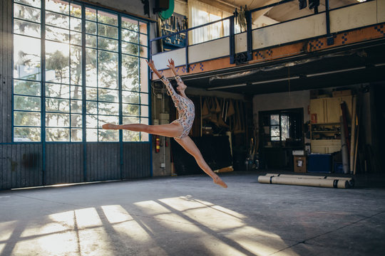 Side view of young slim ballerina in gymnastic suit jumping while practicing dance movements in light spacious studio with large window