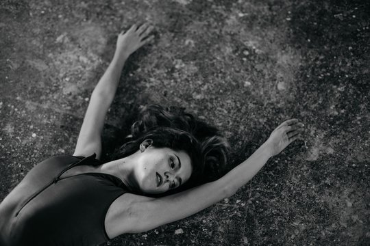 Black and white of alluring relaxed young female in bodysuit lying on shabby ground looking away