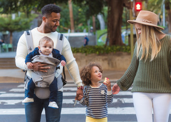 Happy young multiracial parents with little daughter eating apple and cute infant boy crossing road on zebra while walking in city in summer day