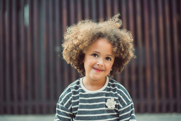 Adorable little girl with curly hair wearing casual striped shirt smiling while standing against blurred wall on street